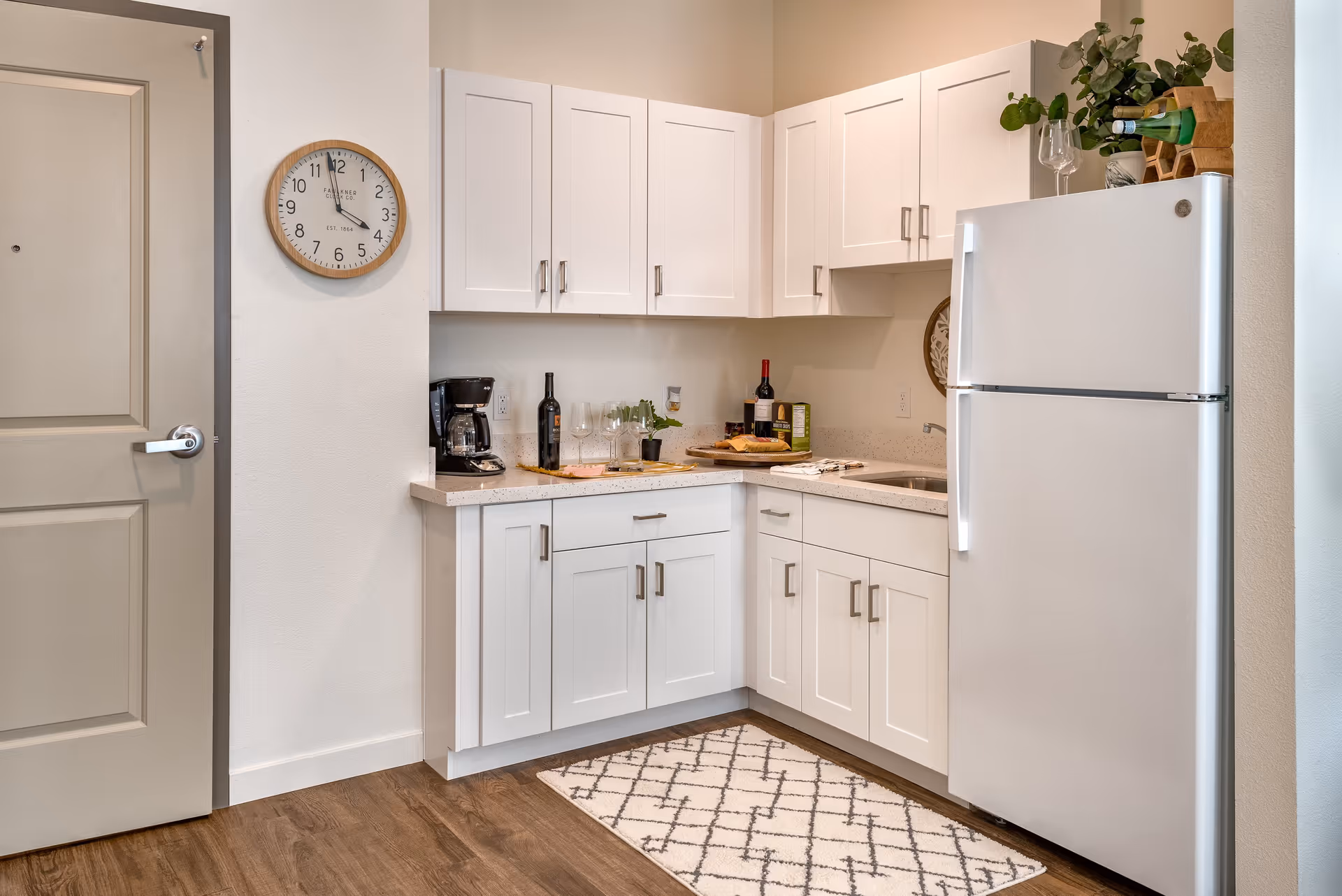 A compact modern kitchenette with white cabinetry, a refrigerator, coffee maker, wine bottles, and a wall clock.