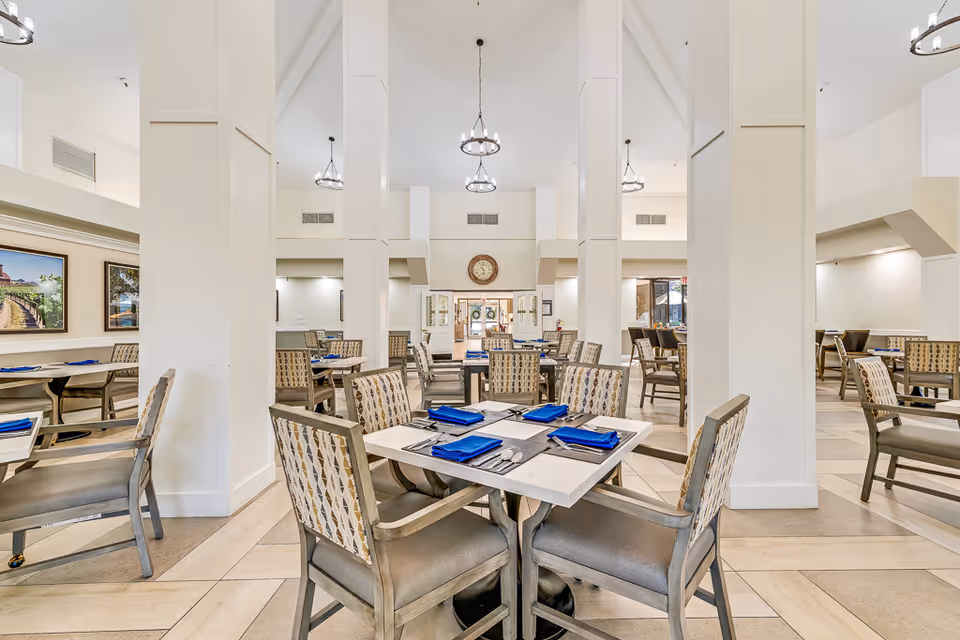 A bright and spacious dining room with multiple tables and chairs arranged neatly. Each table is set with blue napkins, placemats, and silverware. The room features high ceilings with hanging light fixtures, white walls, and framed artwork. There are large pillars dividing the space and a clock mounted on the far wall above a doorway.