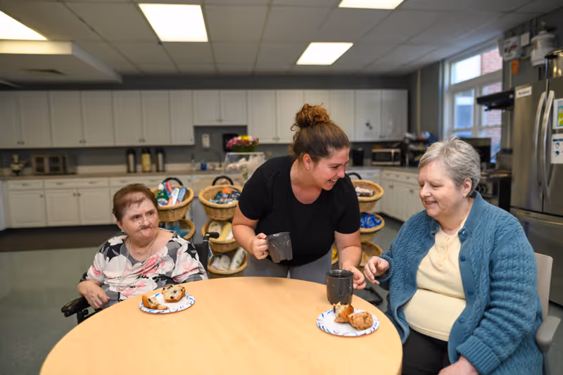 A caregiver serving coffee to two elderly women seated at a round table in a kitchen area. The women have plates with muffins in front of them. The kitchen has white cabinets, a refrigerator, and baskets with snacks in the background.