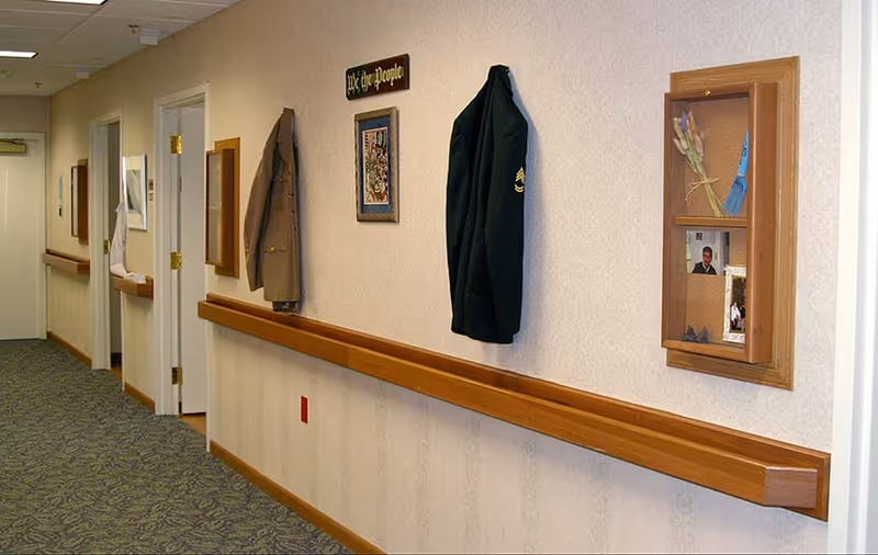 A hallway in a senior living facility with patterned carpet and light-colored walls. Wooden handrails run along the wall, which is decorated with framed pictures, a small display case, and two military jackets hanging on hooks. Several doors lead to rooms along the hallway.