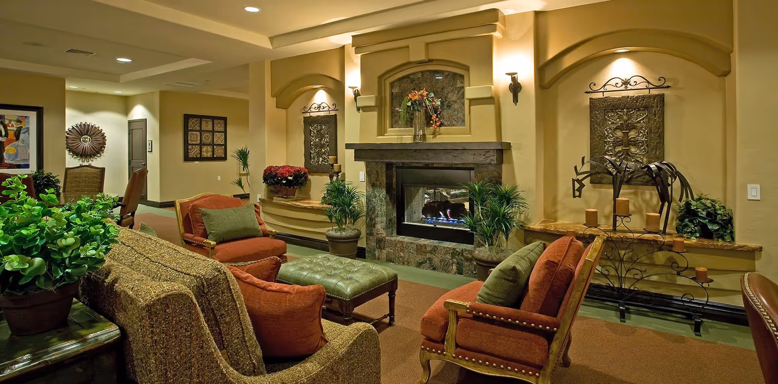 A cozy living room area with a stone fireplace in the center, surrounded by comfortable upholstered chairs and a sofa with cushions. There are decorative plants, wall art, and candle holders enhancing the warm ambiance of the room.