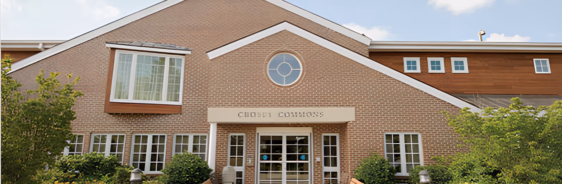 Exterior view of a brick building with a peaked roof and multiple windows. The building has a sign above the entrance that reads 'CHOICE COMMONS'. There are green bushes and trees in front of the building under a clear sky.
