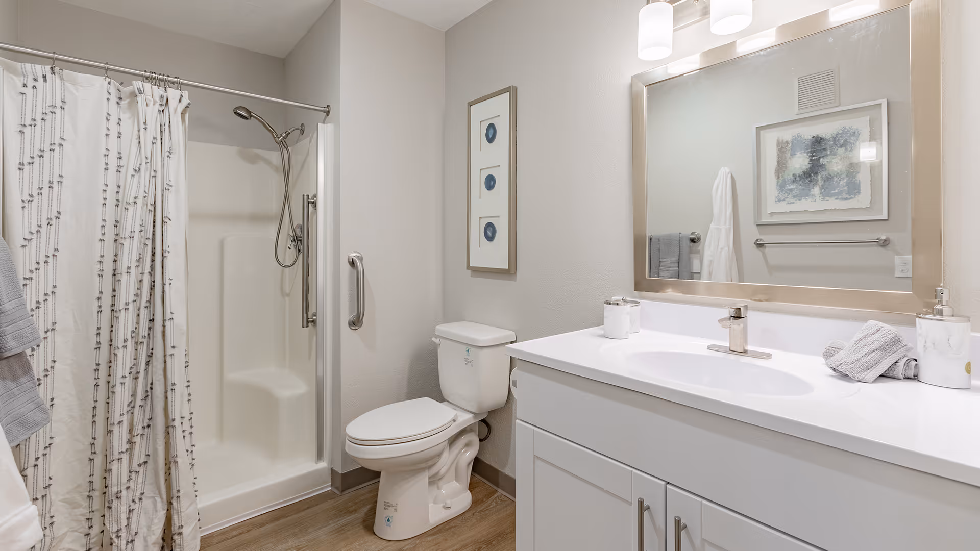 A clean and modern bathroom featuring a white toilet, a shower with a white curtain, a white vanity with a sink, a large mirror above the sink, and framed artwork on the wall. The floor has wood-like tiles and the walls are painted light gray.