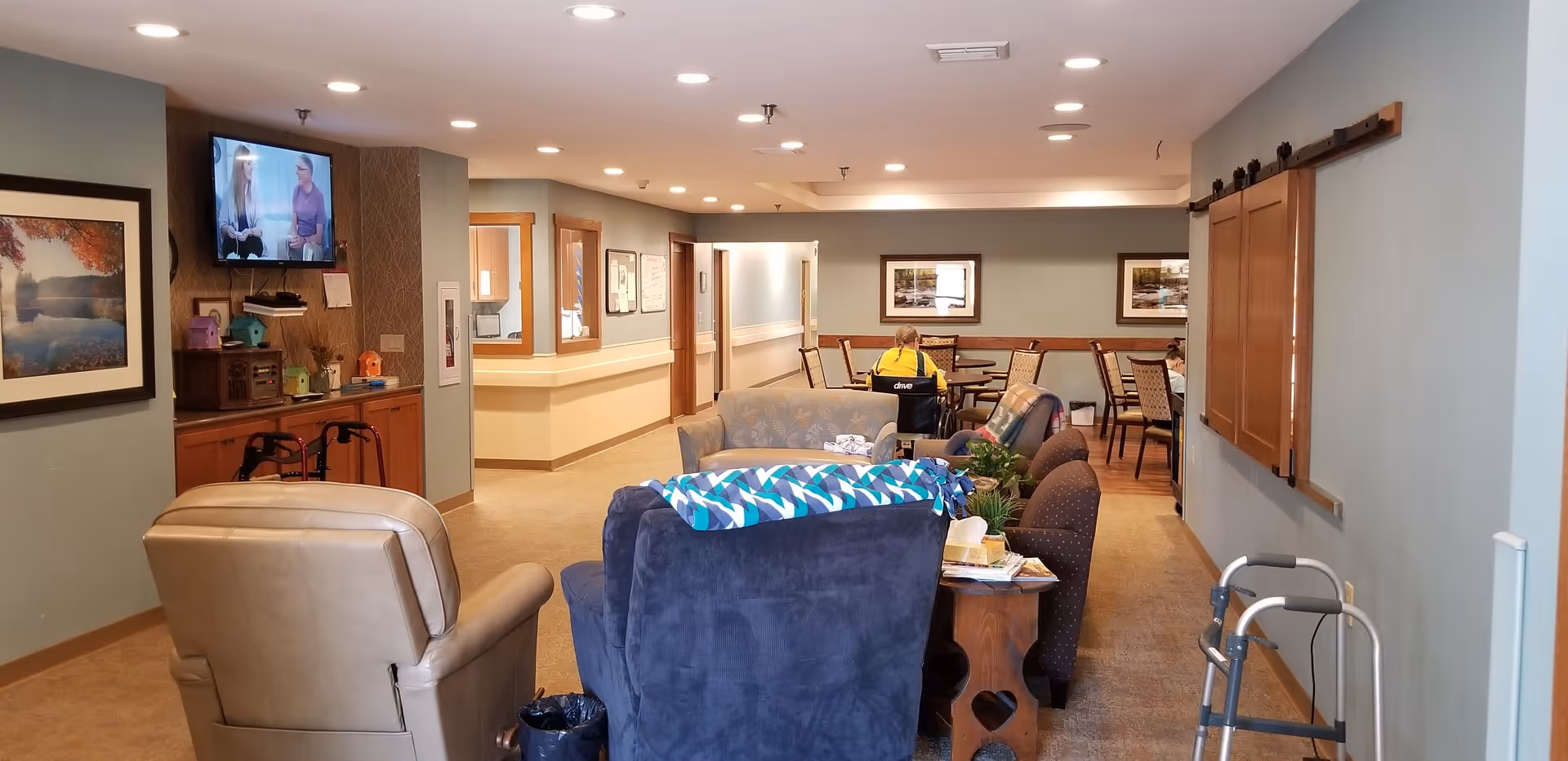 A cozy common area in a senior living facility with several armchairs and sofas arranged around a small table. A television is mounted on the wall to the left, and there are framed pictures on the walls. In the background, a person in a yellow shirt is seated at a dining table. A walker is visible on the right side of the image.