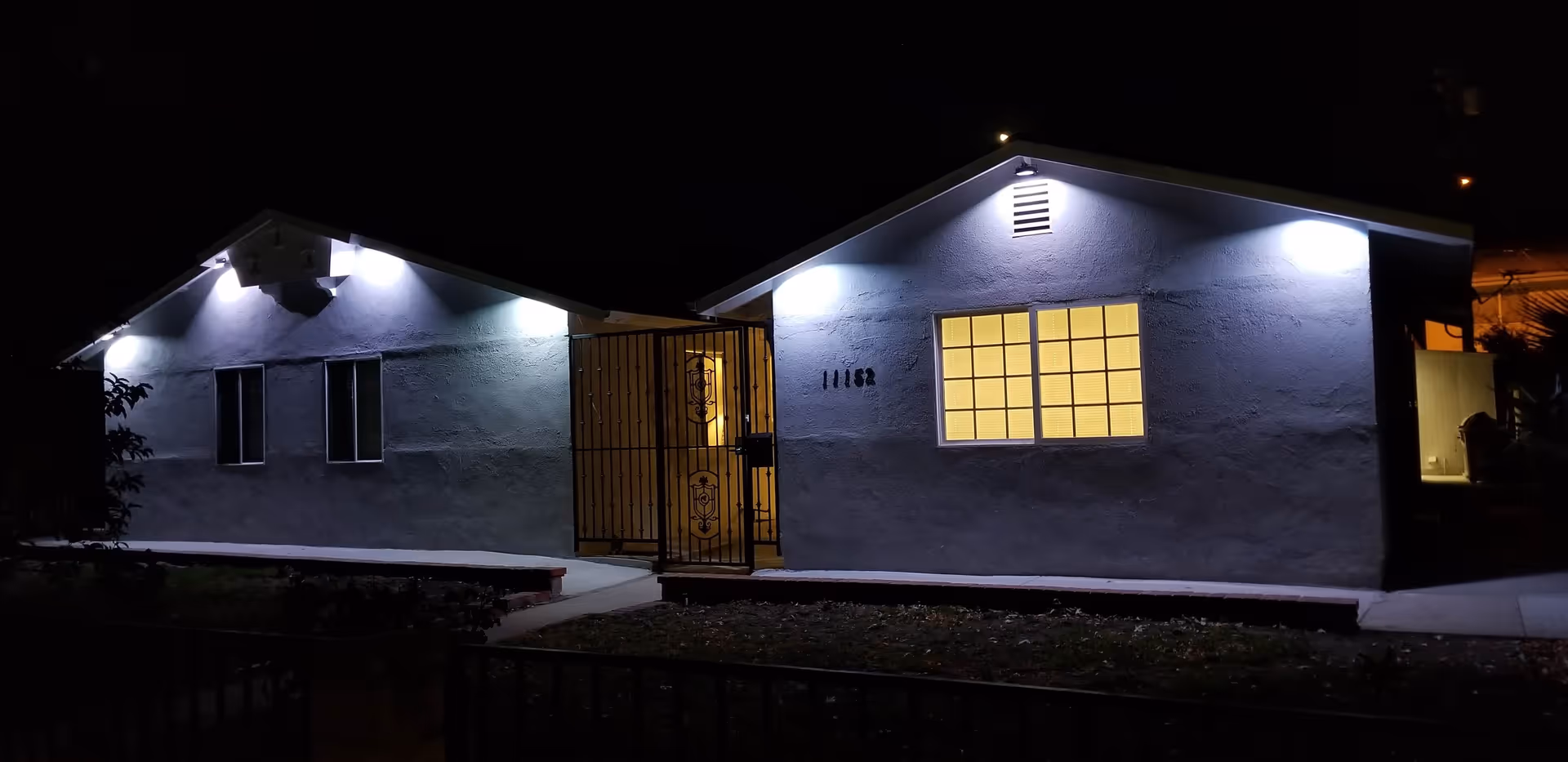 Front exterior of a single-story care home at night with lit windows, wall-mounted lights, and a gated entry.