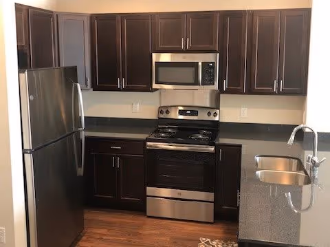 Modern kitchen with dark wood cabinets, stainless steel refrigerator, oven, microwave, and a double sink with a chrome faucet. The countertops are dark, and the floor is wooden.
