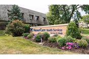 Outdoor view of a landscaped area with a sign that reads 'ManorCare Health Services Nursing & Rehab' in front of a building with brown shingles and surrounded by trees and bushes.