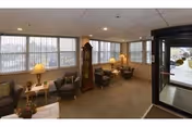 A cozy interior common area with several armchairs arranged around small tables, a grandfather clock, and large windows letting in natural light. There are lamps on the tables and a glass door leading outside.