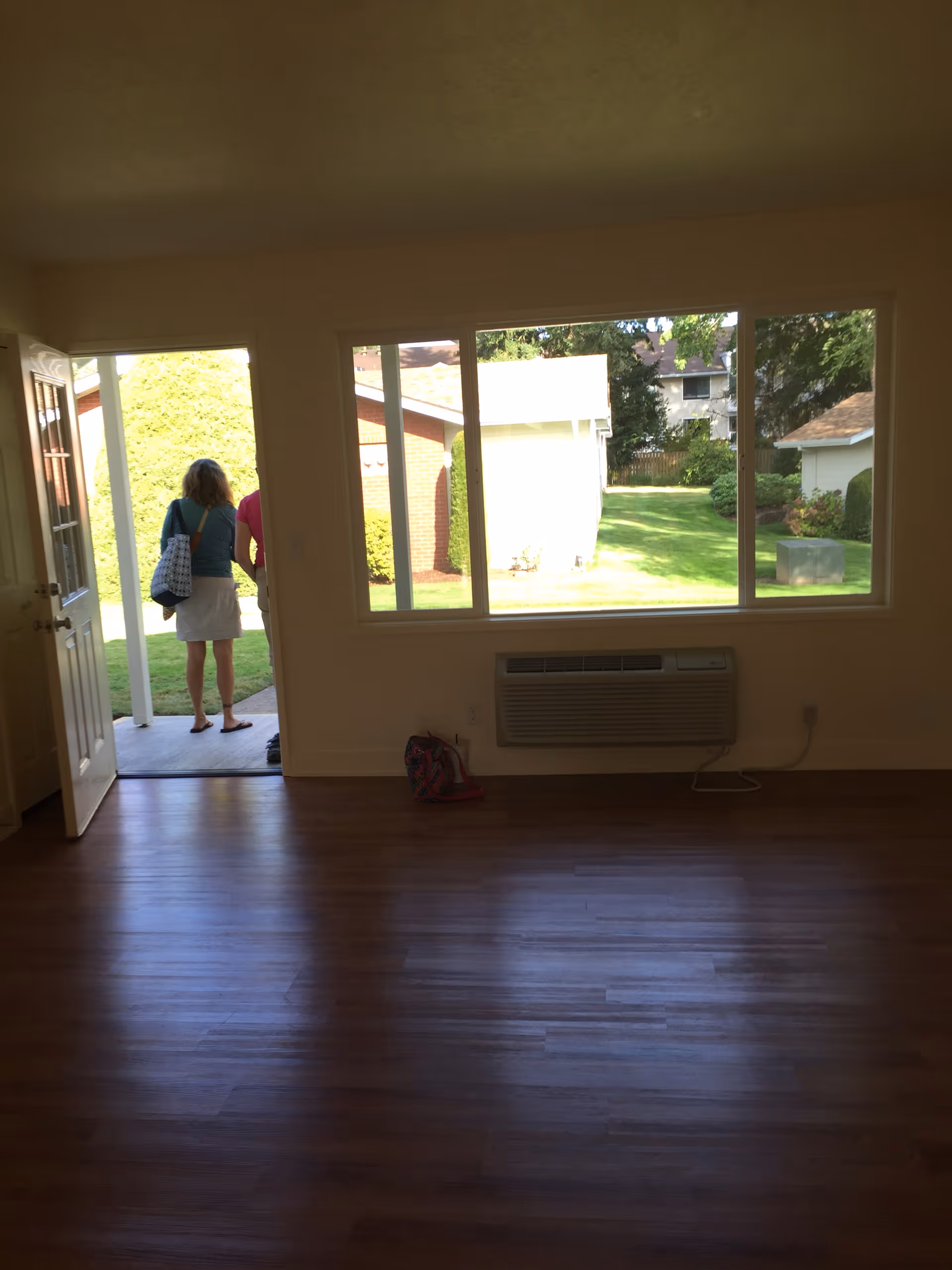 Empty living room with hardwood floors, a large window and wall air conditioner facing a green yard, and two people standing outside at the open door.