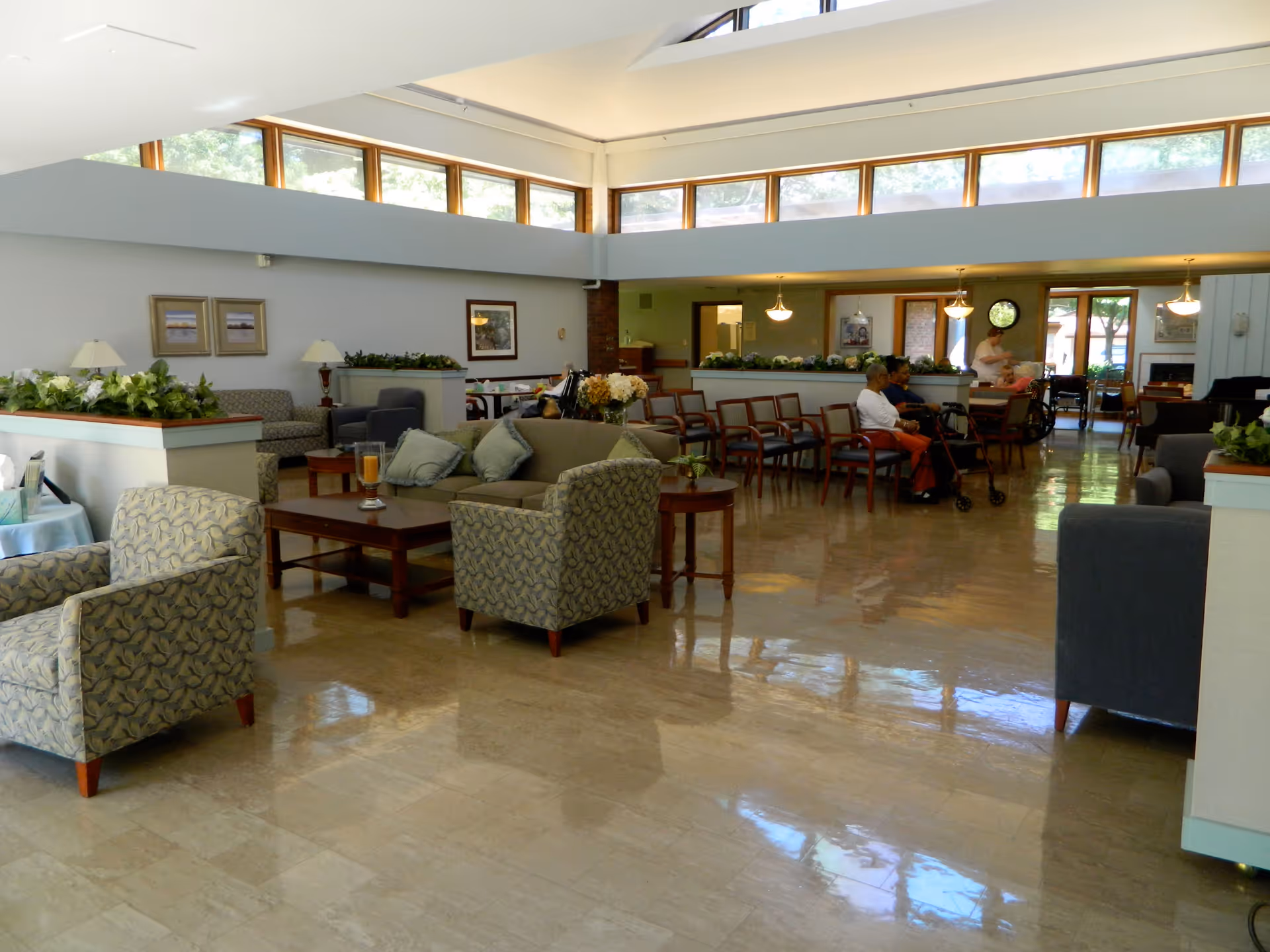A spacious and well-lit common area in a senior living facility with multiple seating arrangements including armchairs and sofas. The room has large windows near the ceiling allowing natural light to fill the space. Several people are seated in the background, some using walkers. The floor is shiny and reflective, and there are plants and framed pictures decorating the room.