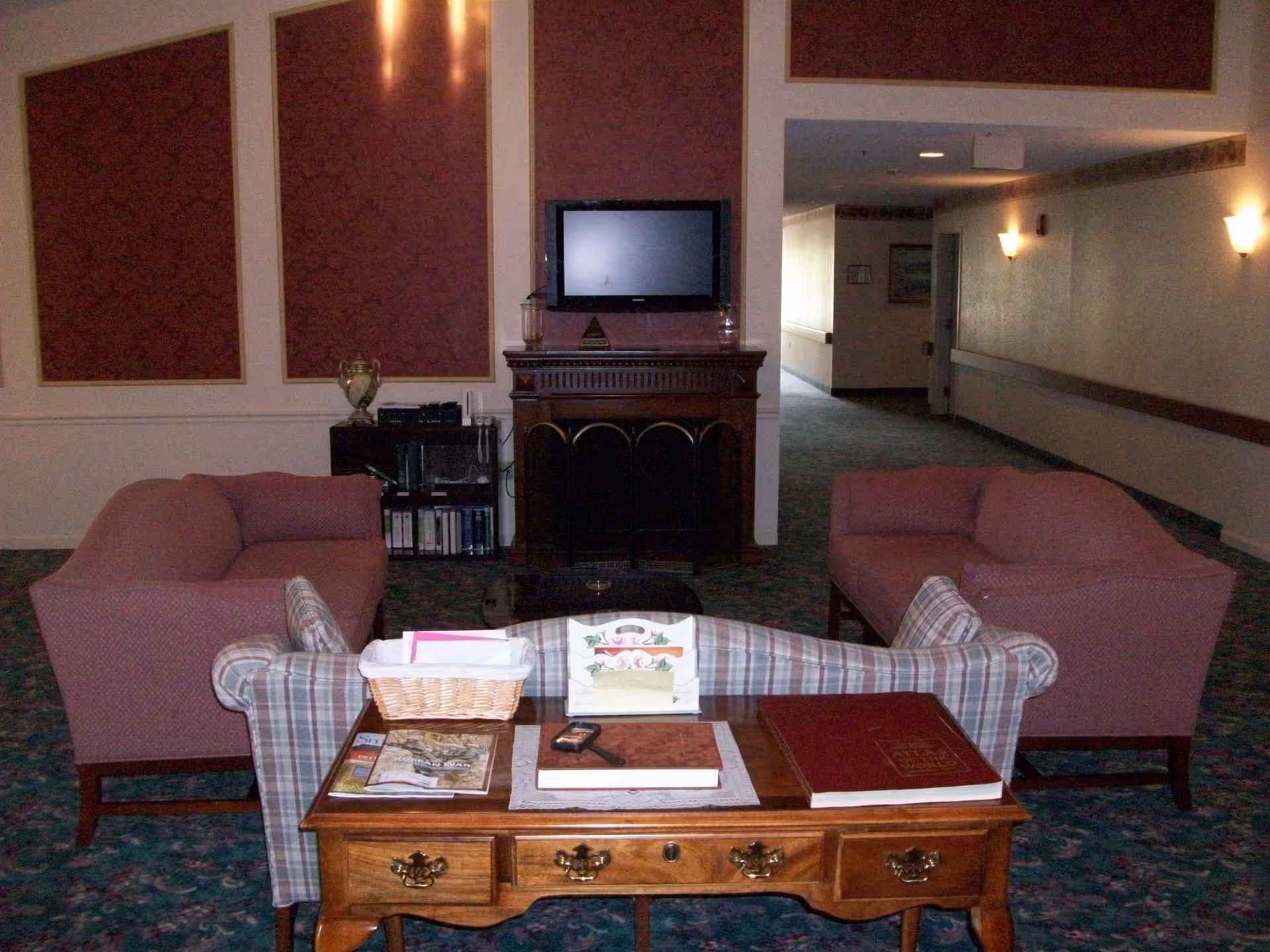 A cozy living room area with two pink upholstered armchairs and a plaid sofa arranged around a wooden coffee table. The table holds books, a basket, and a remote control. A flat-screen TV is mounted above a decorative fireplace against a wall with three large framed panels. The room has patterned carpet and soft wall lighting, with a hallway visible in the background.