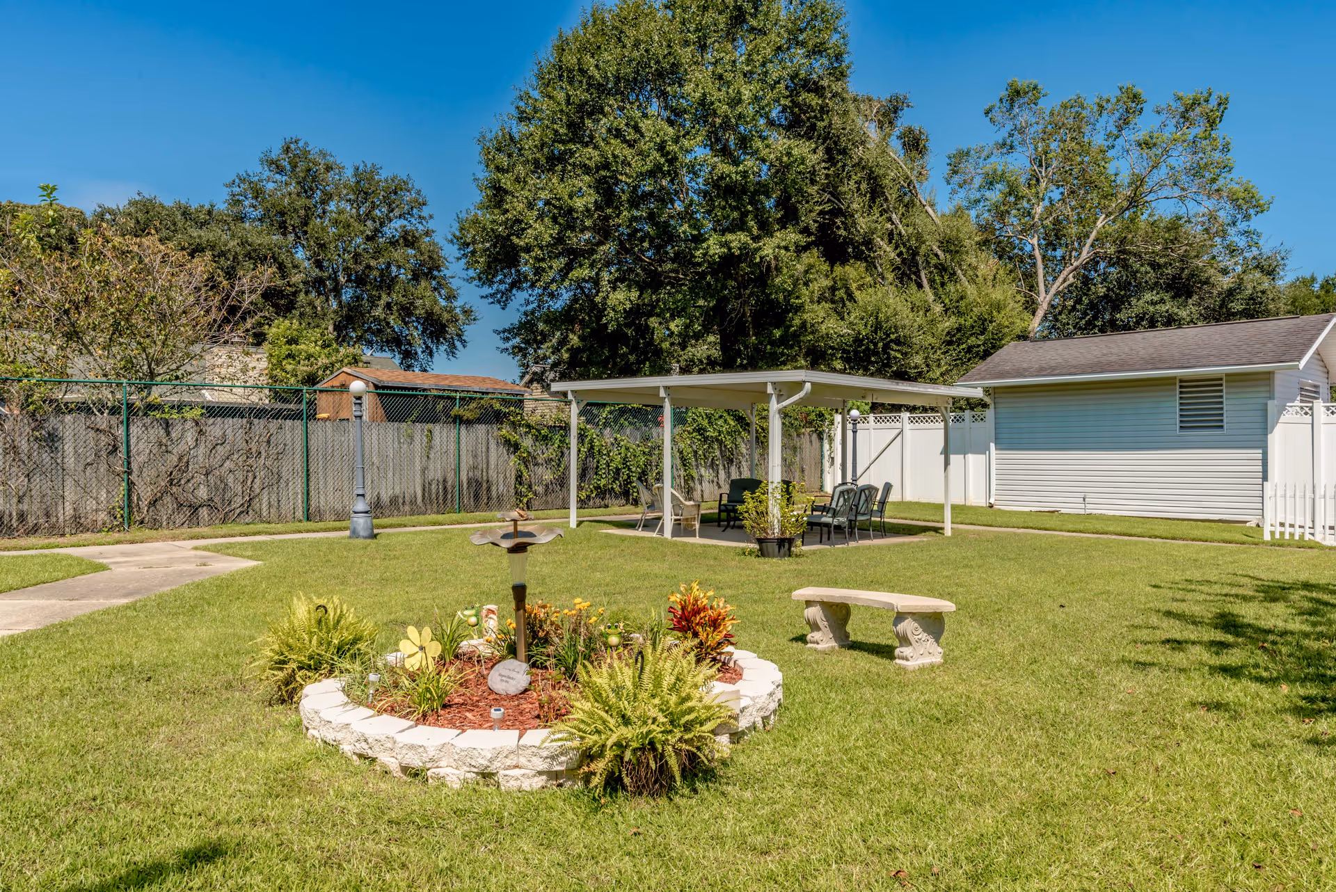 Grassy outdoor courtyard with a covered seating area, a decorative flower bed, and a stone bench.