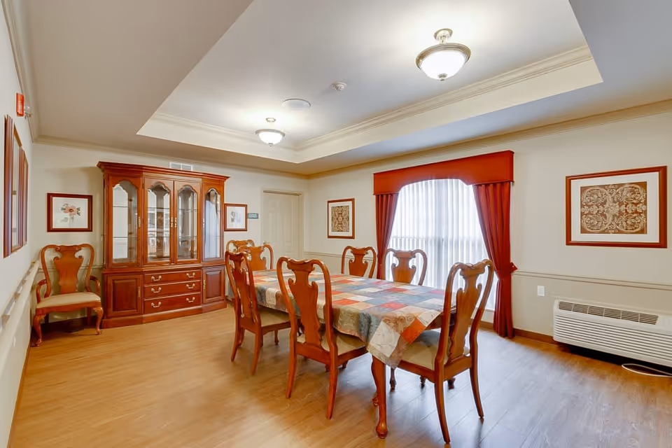 A dining room with a wooden dining table covered with a colorful patchwork tablecloth and eight matching wooden chairs. The room features a wooden china cabinet with glass doors, framed artwork on the walls, a large window with red curtains, and ceiling lights. The floor is wooden, and there is a wall-mounted heater below the window.