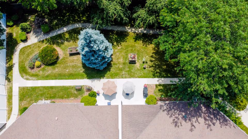 Aerial view of a senior living facility's outdoor garden area with green grass, trees, shrubs, two patio umbrellas, and a concrete walkway. The roof of the building is visible at the bottom of the image.