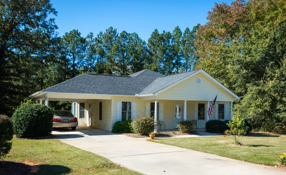 Single-story light yellow house with a carport, driveway and American flag surrounded by trees and lawn.