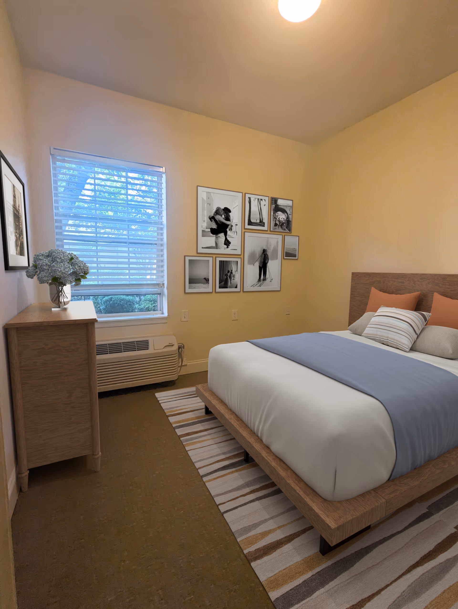 Bedroom with a low wooden bed, striped rug, dresser by a window with blinds and a gallery wall of framed photos.