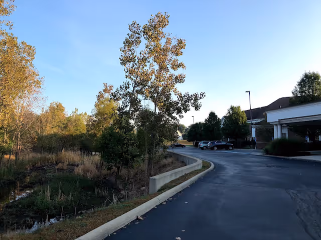 A paved driveway curves alongside a landscaped area with trees and grass, leading to the entrance of a building with a covered drop-off area. Several parked cars are visible near the building under a clear blue sky.
