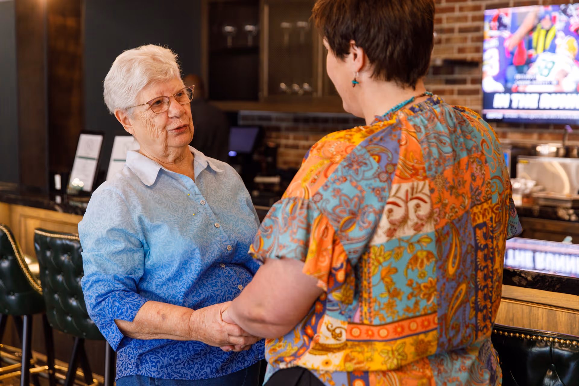 Two women holding hands and engaging in conversation in a cozy indoor setting with a bar counter and green bar stools in the background. One woman has short white hair and glasses, wearing a blue patterned shirt, while the other woman has short brown hair and is wearing a colorful patterned blouse.