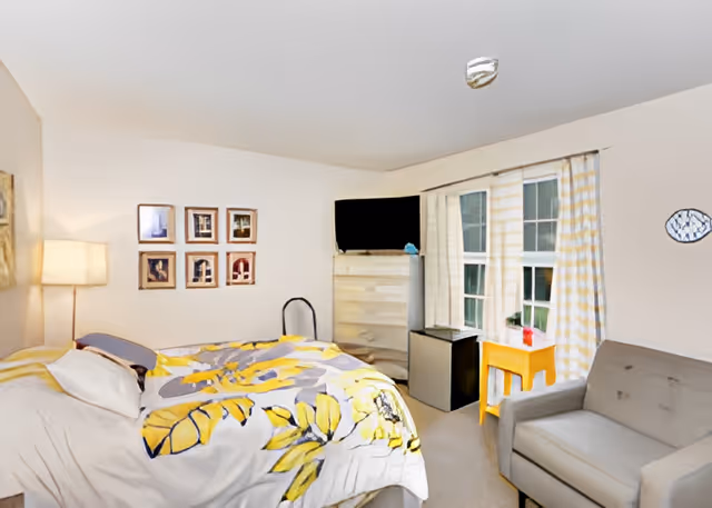 A cozy bedroom with a bed covered in a floral yellow and gray comforter, a floor lamp beside the bed, six framed pictures on the wall, a dresser with a flat-screen TV on top, a small refrigerator next to the dresser, a window with yellow and white checkered curtains, a small yellow table with a plant on it, and a gray armchair.