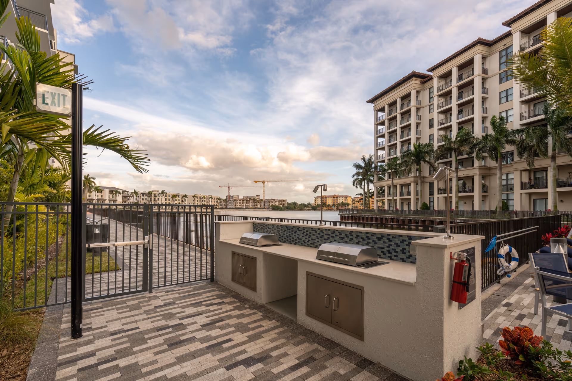 Outdoor area at Overture Doral featuring two built-in stainless steel grills on a tiled countertop with a mosaic tile backsplash. The area is paved with gray and white bricks and enclosed by a black metal fence with an exit sign. In the background, there is a large body of water, palm trees, and multi-story residential buildings under a partly cloudy sky.