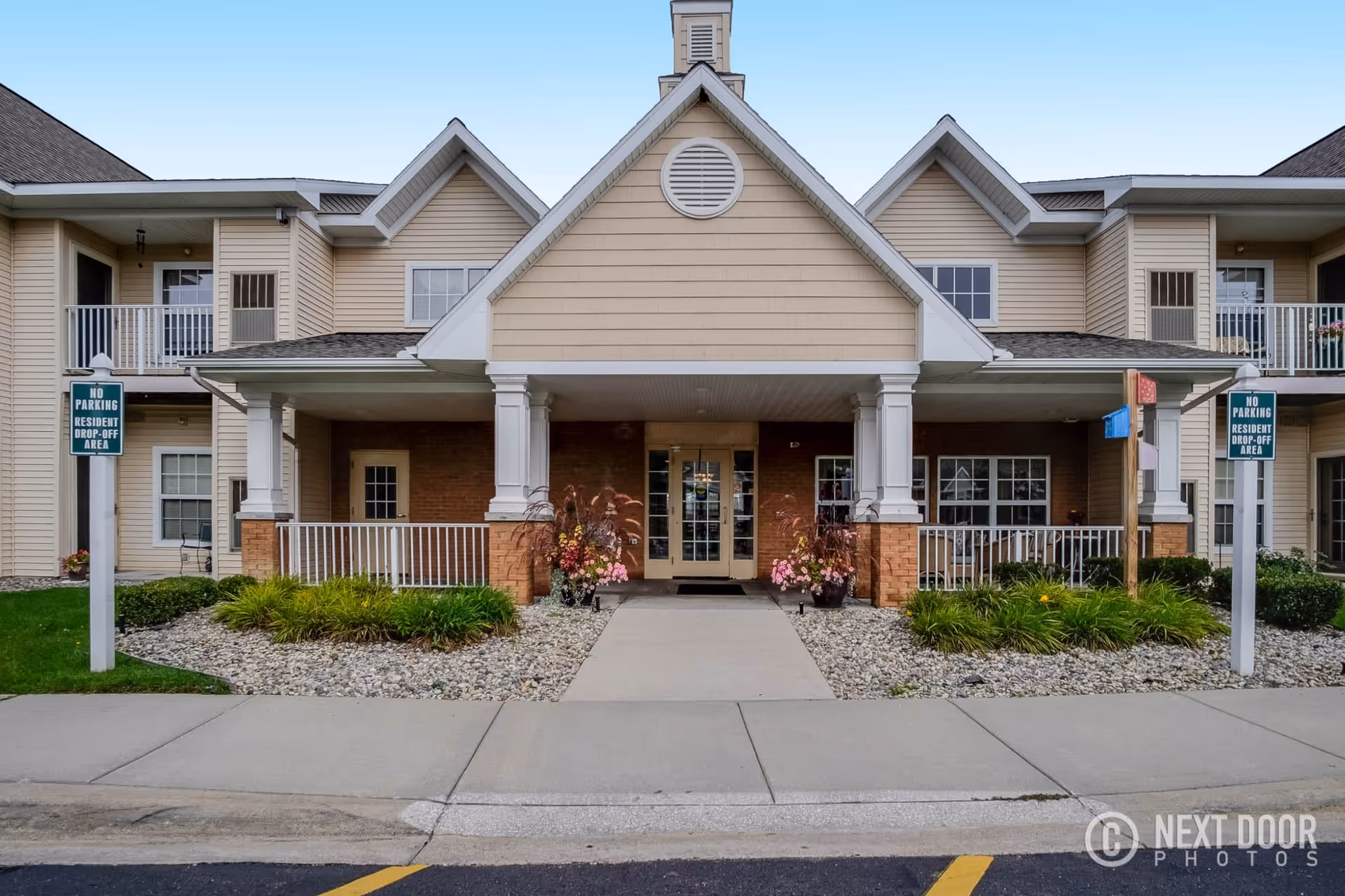 Front exterior view of Devonshire Retirement Village showing a two-story building with beige siding and brick accents. The entrance has a covered porch supported by white columns, with flower pots on either side of the door. There are balconies on the second floor and signs indicating no parking in the resident drop-off area.