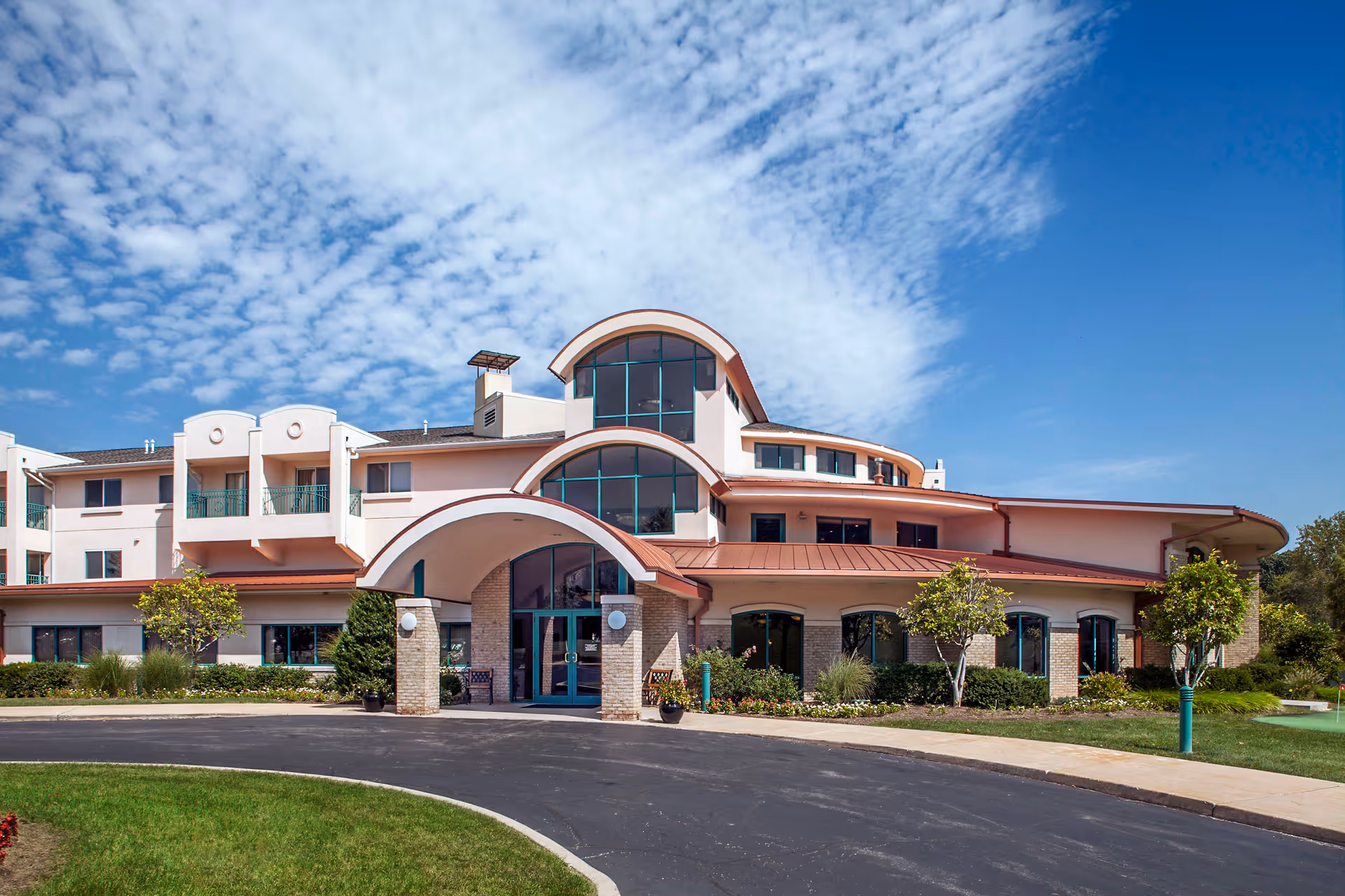 Front exterior view of a multi-story senior living facility building with arched windows and entrance, surrounded by landscaped greenery and a clear blue sky with scattered clouds.