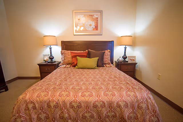 A neatly made bed with a patterned bedspread and decorative pillows flanked by matching nightstands and lamps, with framed floral artwork above the headboard.
