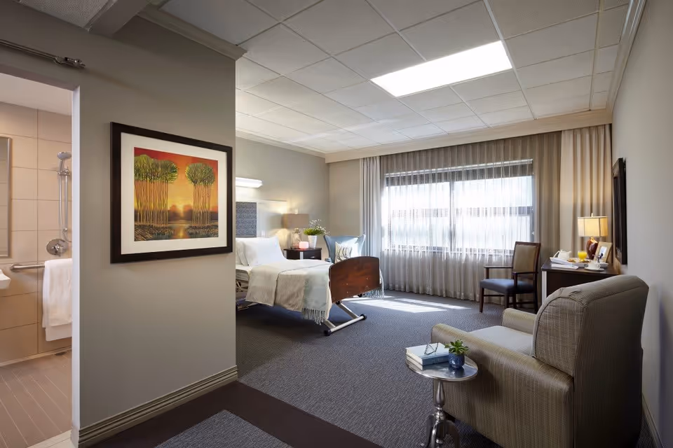 A well-lit, comfortable room in a senior living facility featuring a hospital-style bed with white linens and a light gray blanket. The room includes a cushioned armchair with a small round side table holding books and a small plant. There is a wooden chair and a desk with a lamp, a glass of orange juice, and a teapot. A large window with sheer curtains allows natural light to fill the room. On the left side, a bathroom with tiled walls and a shower is partially visible. A framed artwork depicting trees is hung on the wall near the bathroom entrance.