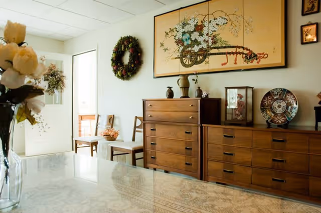 A cozy interior room featuring wooden dressers with decorative items including vases and a display case. A floral wreath hangs on the wall next to a door with a window. Above the dressers is a large framed artwork depicting a cart filled with flowers. Two wooden chairs are positioned near the door, and a vase with white flowers is visible in the foreground on a table with a patterned tablecloth.