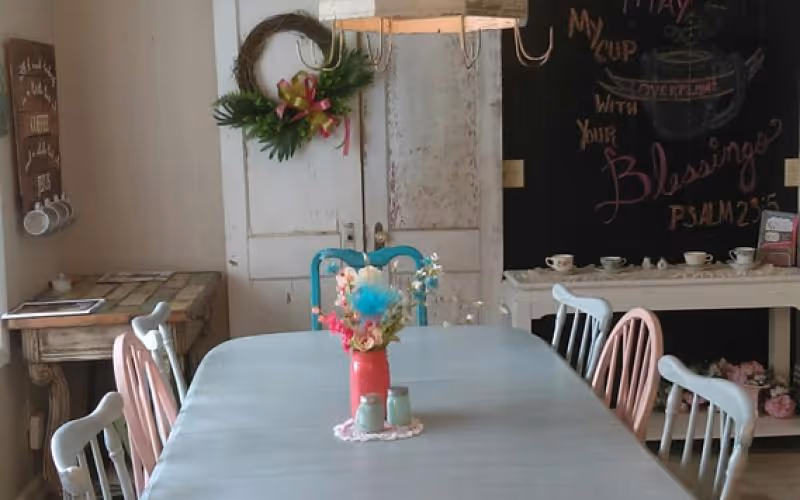 A cozy dining room with a long light blue table surrounded by mismatched pastel-colored chairs. A red vase with colorful flowers and two small jars sit on a doily in the center of the table. In the background, there is a rustic white cabinet with a wreath hanging on it, a small wooden table with cups hanging on the wall, and a blackboard with a handwritten inspirational quote from Psalm 23:5.