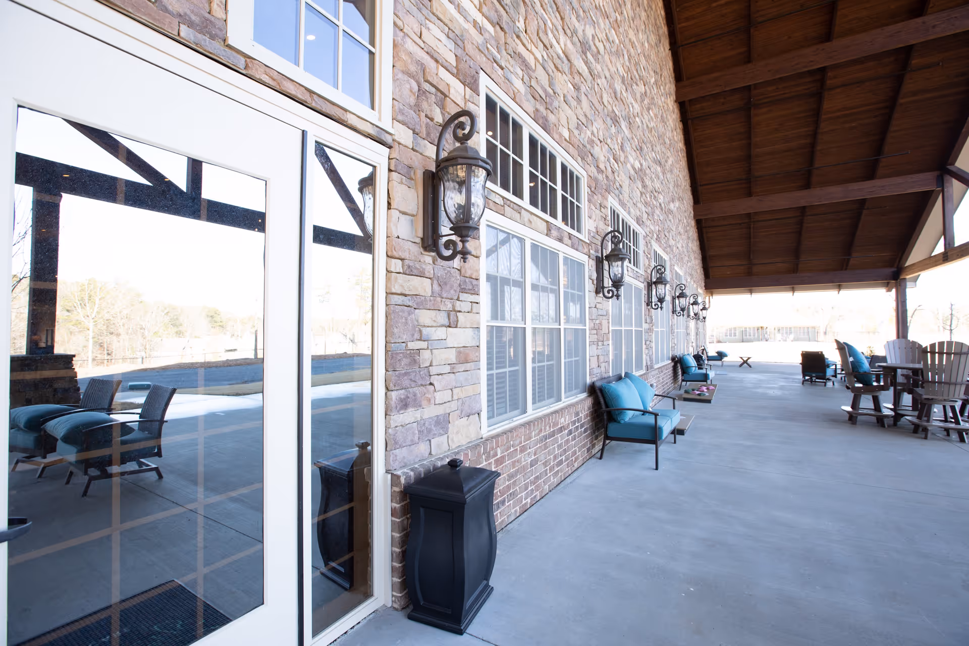 Covered outdoor patio along a stone building wall with windows, decorative wall lanterns, and teal-cushioned seating under a wooden roof.