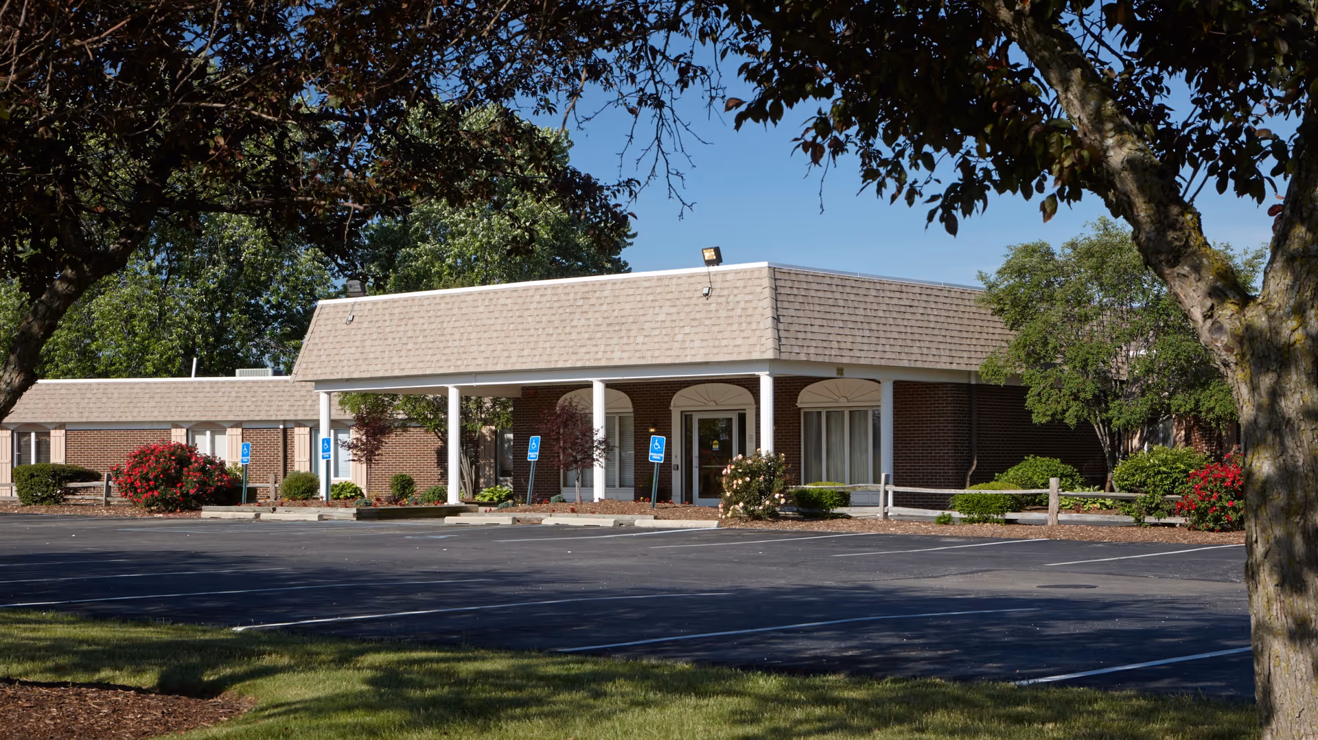 Exterior view of a single-story brick building with a covered entrance supported by white columns. The building is surrounded by trees, bushes, and a parking lot with several handicap parking signs visible. The sky is clear and blue.