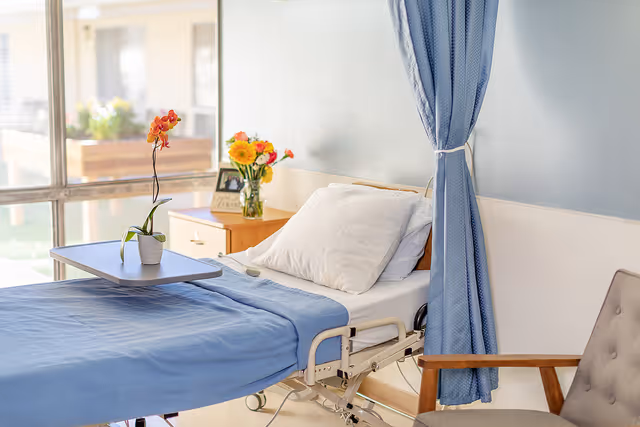 A hospital-style bed with blue bedding and a white pillow in a well-lit room. A small table on wheels is placed over the bed with a potted orchid on it. Behind the bed is a wooden nightstand with a vase of flowers and a framed photo. A blue privacy curtain is partially drawn to the side, and a cushioned chair with wooden armrests is positioned nearby. Large windows let in natural light.