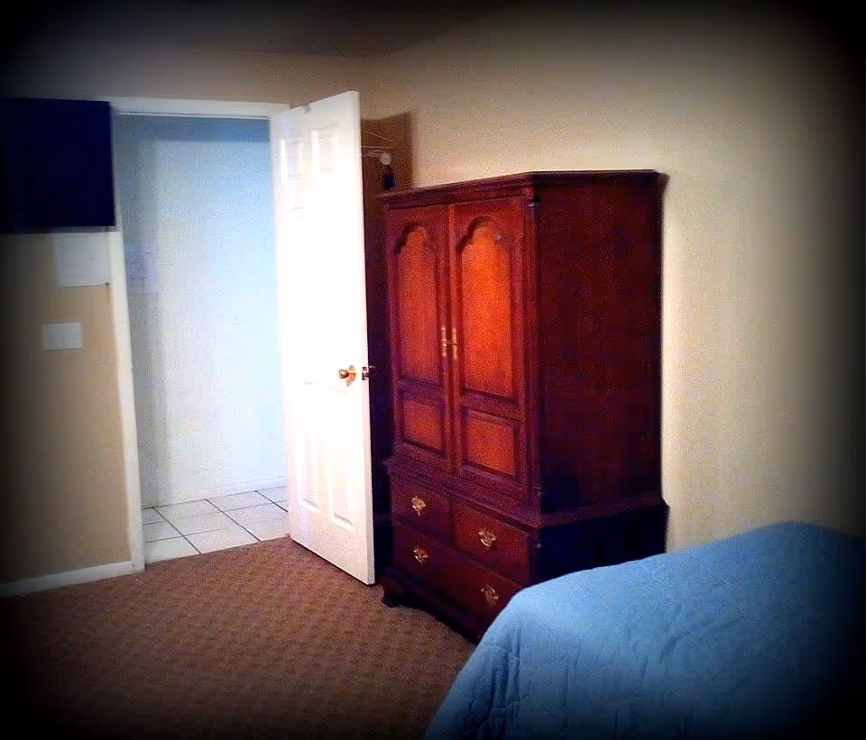 A bedroom with a wooden armoire, a partially open white door leading to a tiled area, beige walls, carpeted floor, and a bed with a blue quilted bedspread visible in the foreground.