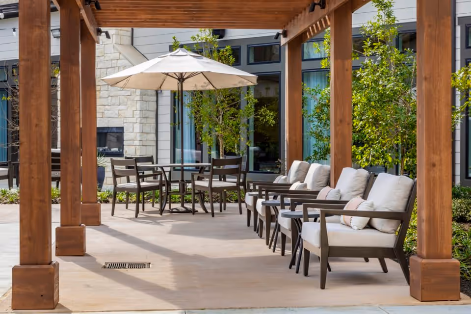 Outdoor patio area at The Preserve at Spring Creek featuring cushioned chairs with pillows arranged under a wooden pergola, a round table with chairs, and a large umbrella. The background shows building windows and greenery.