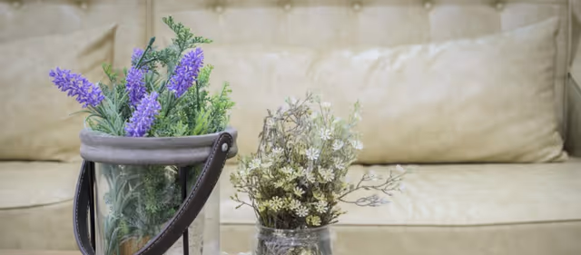 Two small vases with purple and white artificial flowers sit on a table in front of a beige tufted sofa.