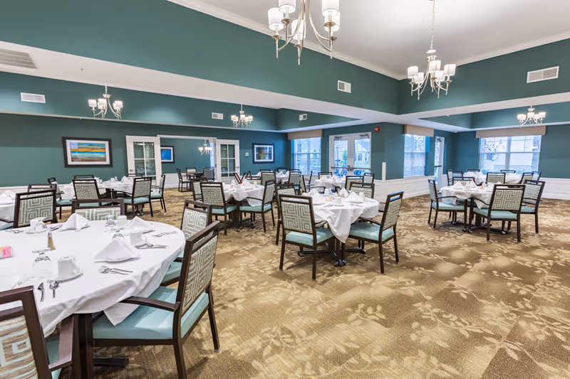 A spacious dining room with multiple round tables covered in white tablecloths, each set with napkins, cups, and silverware. The room has teal walls, large windows letting in natural light, patterned carpet, and several chandeliers hanging from the ceiling. Framed artwork is displayed on the walls.