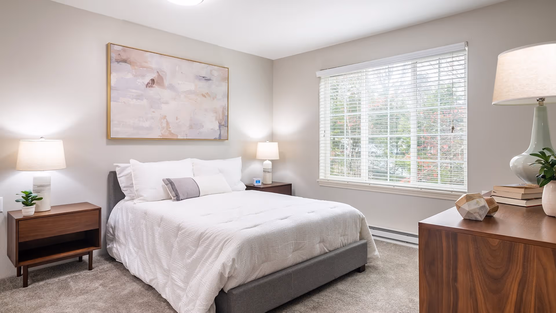 A bright and clean bedroom with a large window covered by white blinds. The room features a neatly made bed with white bedding and multiple pillows, two wooden nightstands each with a lamp and small decorative items, a wooden dresser with a lamp, books, and a plant. A large abstract painting hangs above the bed.