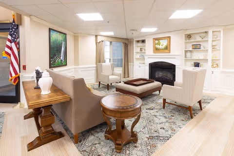 A bright and cozy living room area in a retirement community featuring a beige sofa, two white armchairs, a wooden coffee table, and a round side table. The room has a fireplace with built-in white shelves on either side, decorated with books and ornaments. A large painting hangs above the fireplace, and an American flag stands near the entrance door. The floor is covered with a patterned rug, and the walls are painted light beige with white trim.