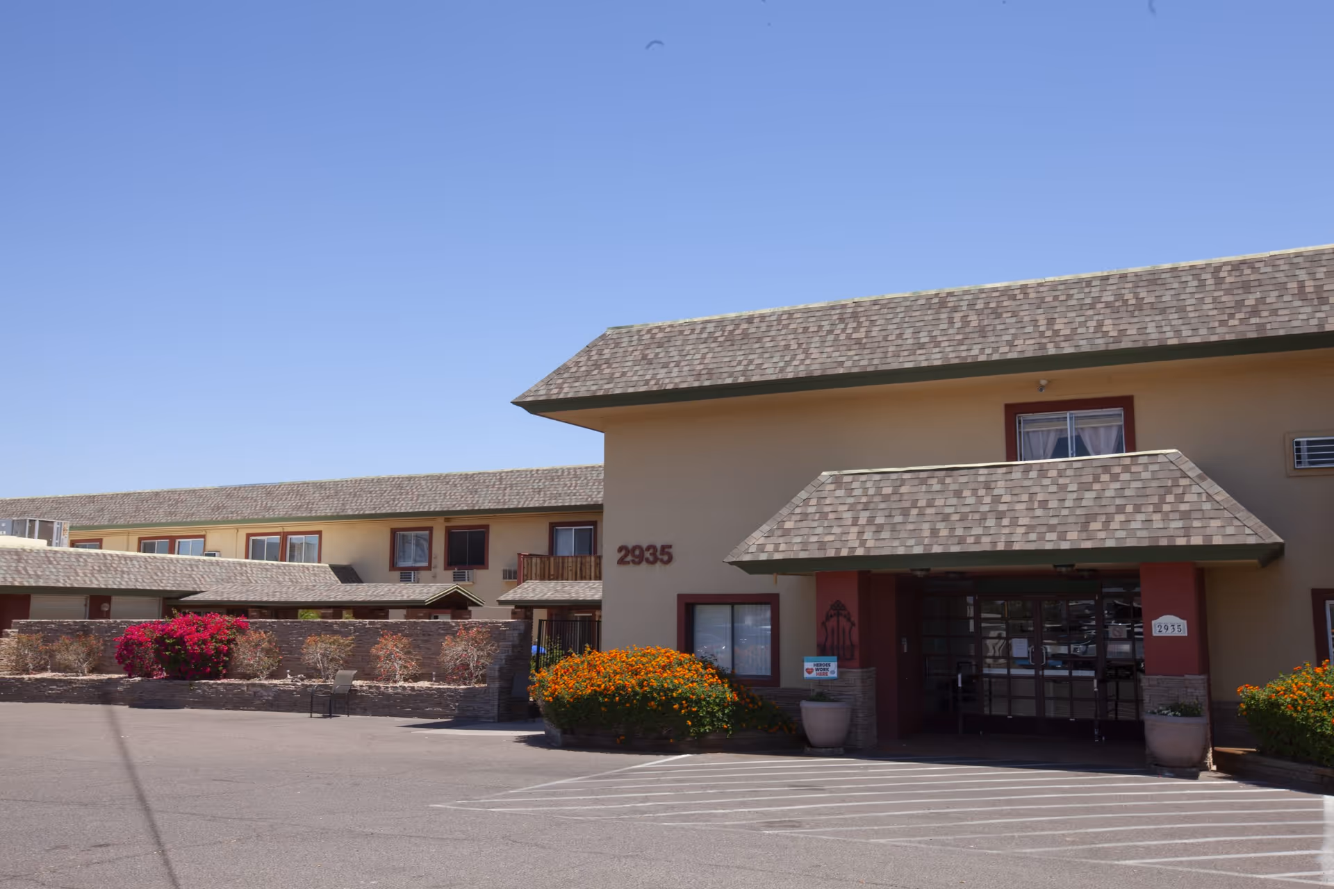 Entrance and front facade of a two-story senior living building with parking lot and flowering landscaping under a clear blue sky.
