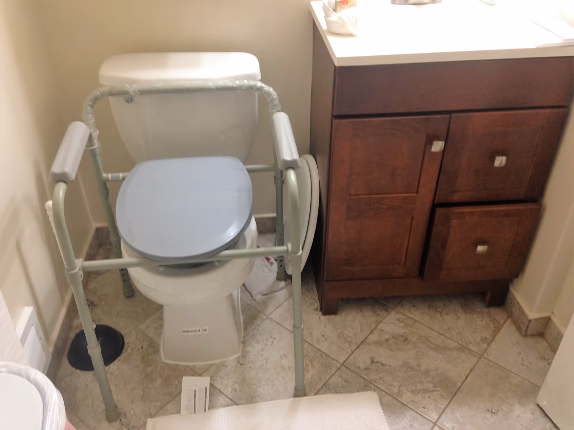 Bathroom with a white toilet equipped with a gray raised toilet seat and safety frame with armrests. Next to the toilet is a wooden vanity cabinet with a white countertop. The floor is tiled with light-colored tiles.