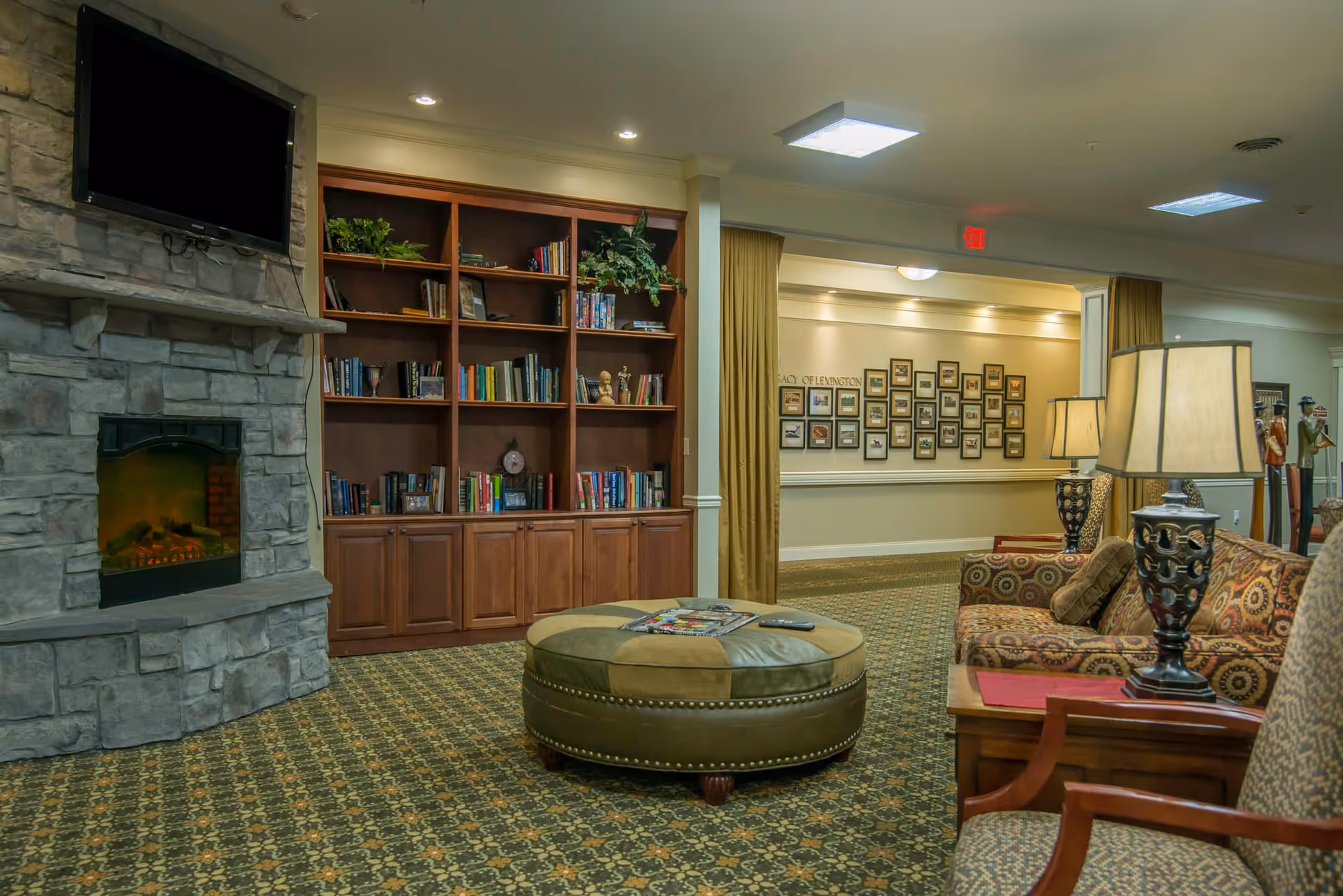 Cozy senior living common room with a stone fireplace, built-in bookcases, round ottoman and upholstered seating with lamps.