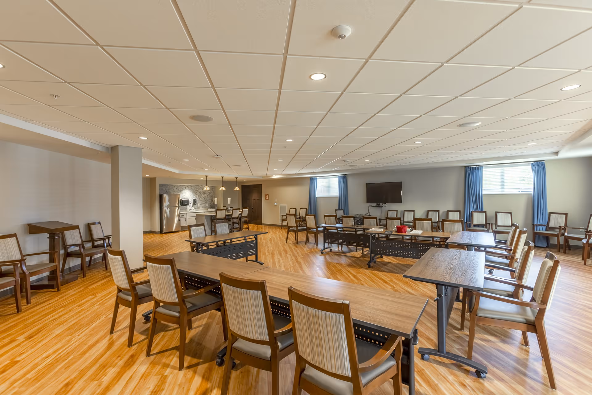 A spacious, well-lit common area with multiple wooden tables and chairs arranged in a U-shape and rows. The room has light wood flooring, a drop ceiling with recessed lighting, two windows with blue curtains, a wall-mounted TV, and a kitchenette area with a refrigerator and pendant lights in the background.