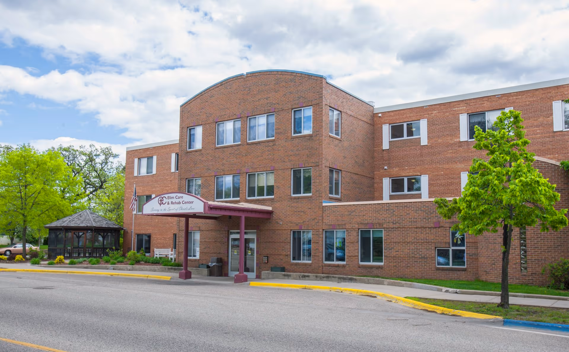Exterior view of a three-story brick building with multiple windows, a covered entrance with a sign reading 'Elim Care & Rehab Center', a small gazebo to the left, and a tree on the right side. The sky is partly cloudy.