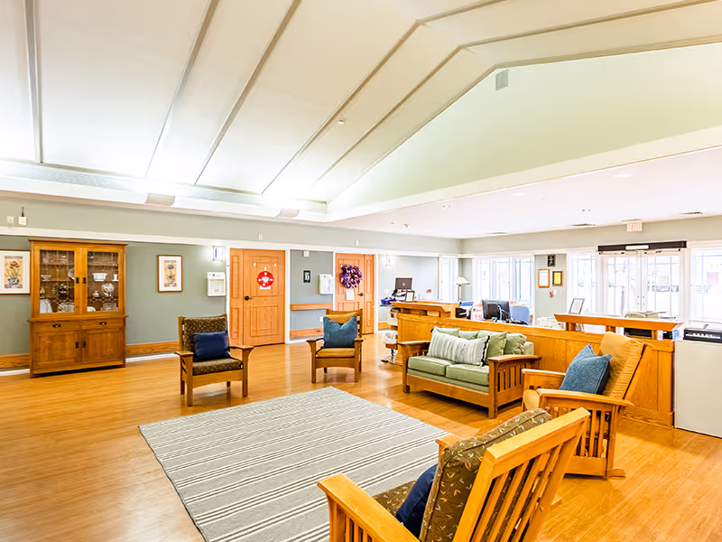 A bright and spacious common area in a senior living facility featuring wooden furniture including chairs and a sofa with cushions, a striped area rug, a wooden cabinet with glass doors displaying decorative items, and a reception desk in the background. The room has light-colored walls, wooden flooring, and large windows allowing natural light to fill the space.