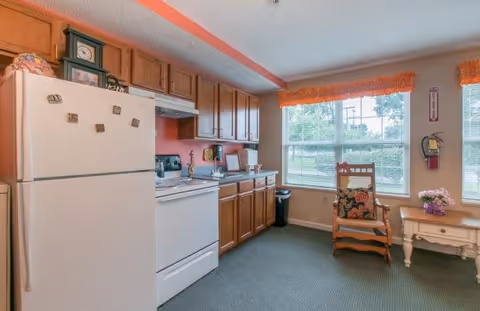 Interior view of a kitchen area in a senior living facility with wooden cabinets, a white refrigerator, and a white stove. There is a window with orange valances letting in natural light. A wooden rocking chair with a cushion and a small side table with a flower pot are placed near the window.