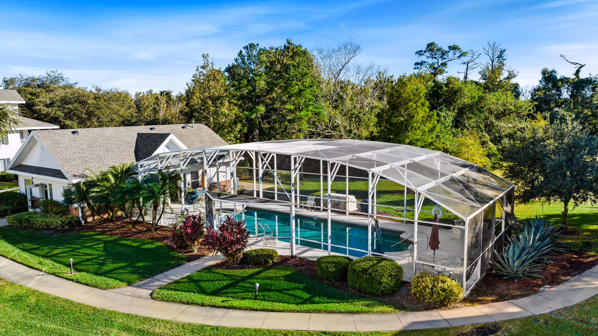 Outdoor view of a senior living community featuring a screened-in swimming pool with lounge chairs and an umbrella inside the enclosure. The pool area is surrounded by well-maintained green lawns, bushes, and trees, with a building visible in the background under a clear blue sky.