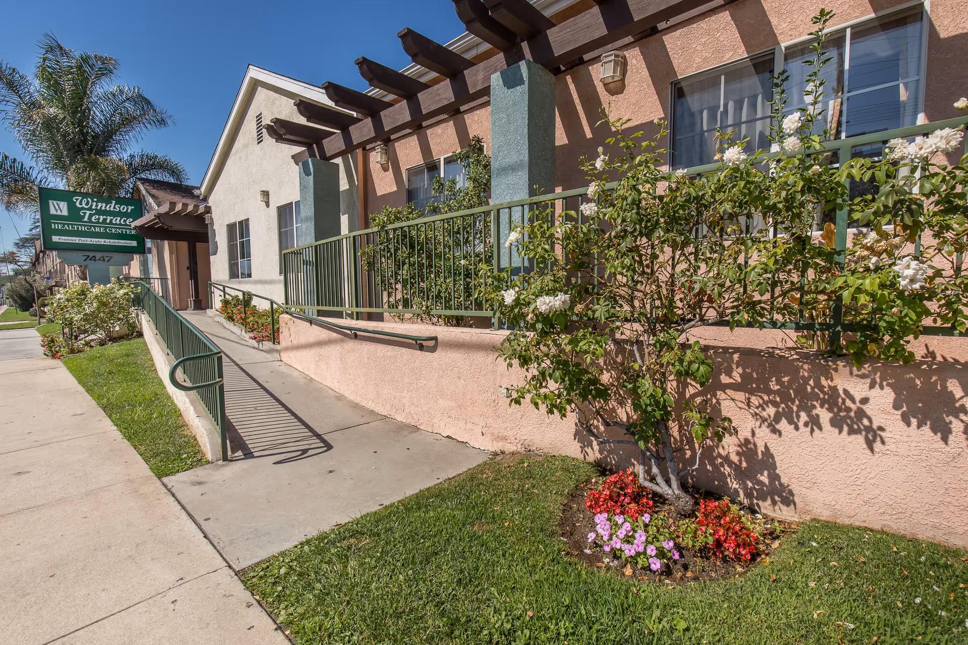 Exterior view of Windsor Terrace Healthcare Center showing a beige and pink building with green railings and a ramp leading to the entrance. There are flowering bushes and a small garden with colorful flowers along the side of the building under a clear blue sky.