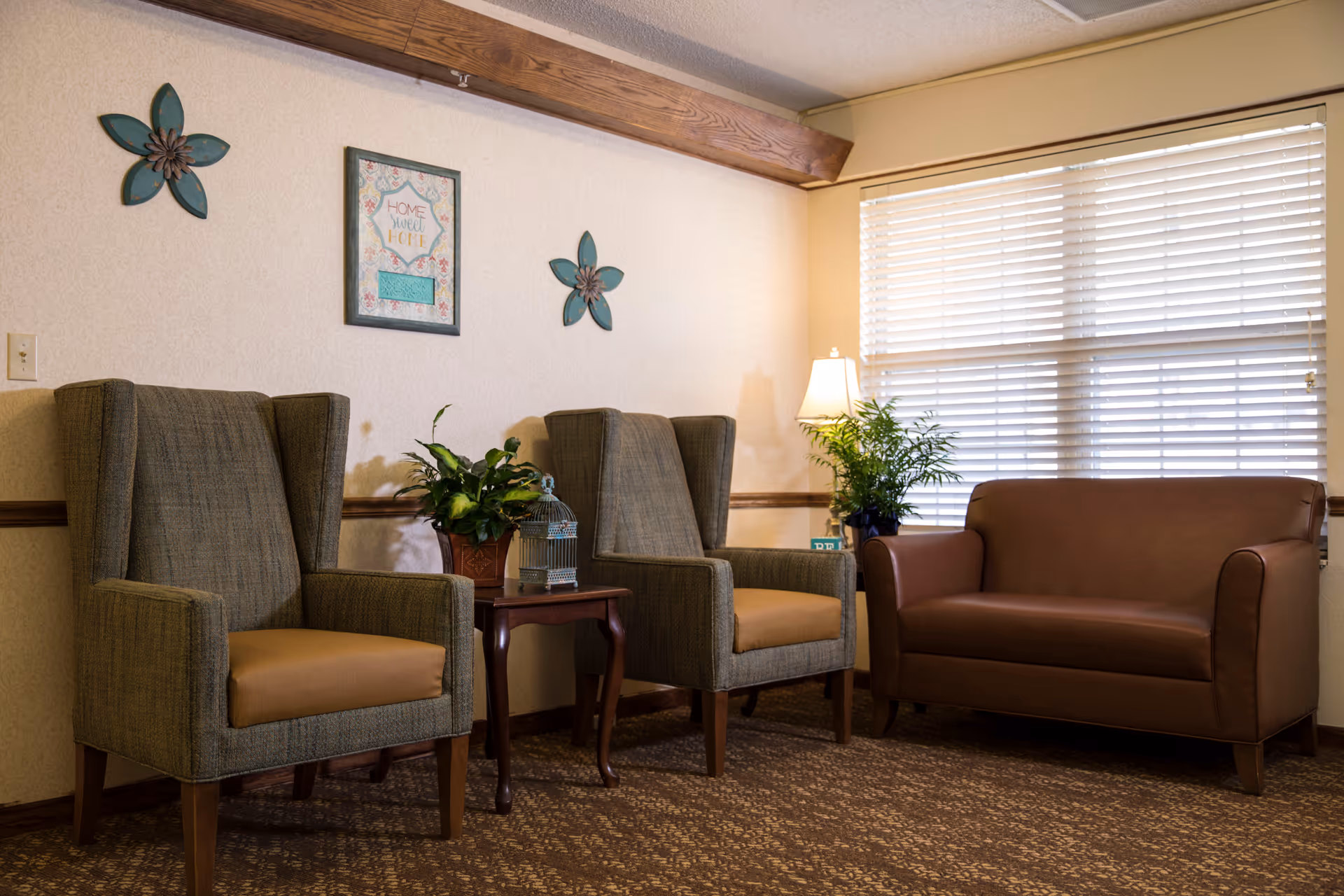 A cozy seating area with two upholstered armchairs, a leather loveseat, a side table with plants and a lamp, and wall decorations by a window with blinds.
