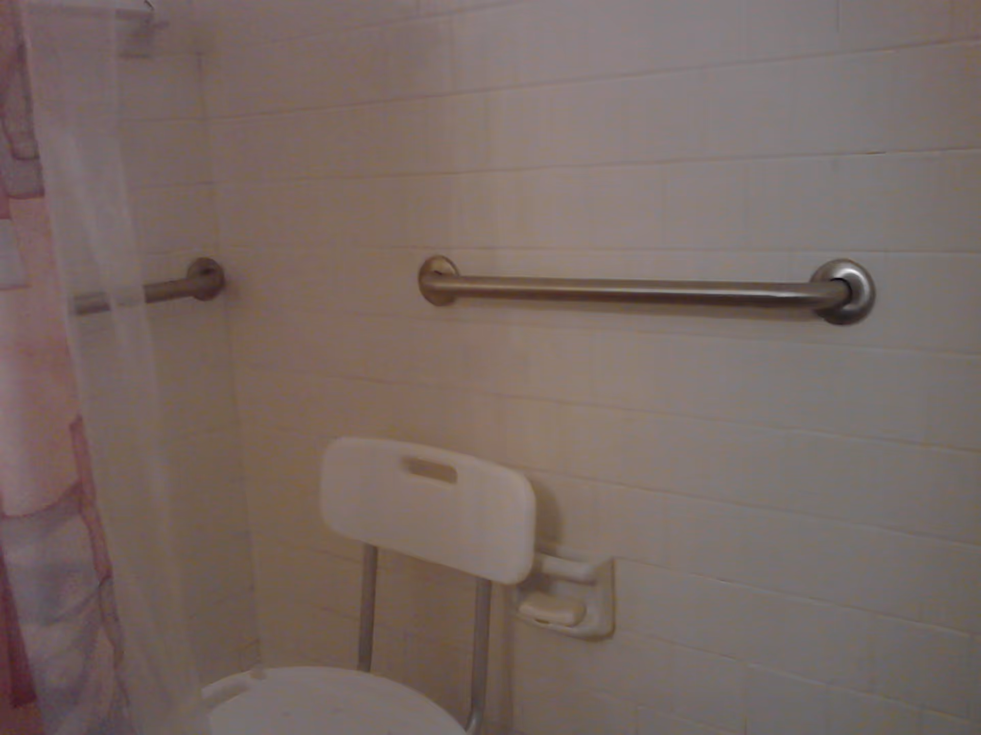 A bathroom shower area with white tiled walls, a white shower chair, a metal grab bar on the wall, and a partially visible shower curtain with pink and purple patterns.